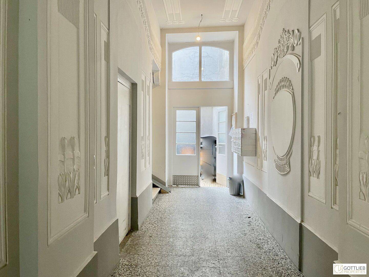 Langer, heller Flur in einem Altbau mit hohen Decken, Stuckelementen und einem Fenster am Ende.