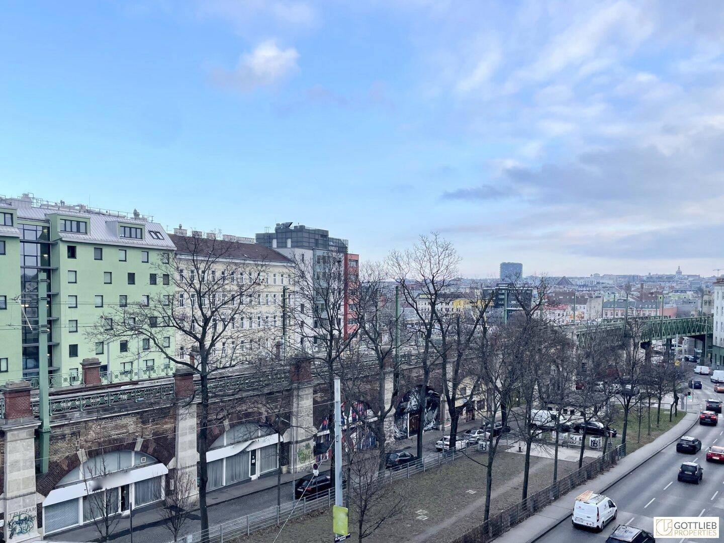 Blick über eine Stadtstraße mit Gebäuden, Bäumen und einem historischen Viadukt unter bewölktem Himmel.
