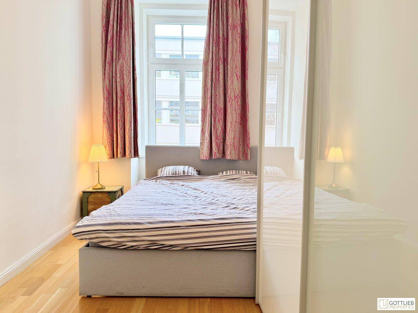 Schlafzimmer mit Doppelbett, Holzboden und zwei Fenstern mit roten Vorhängen.