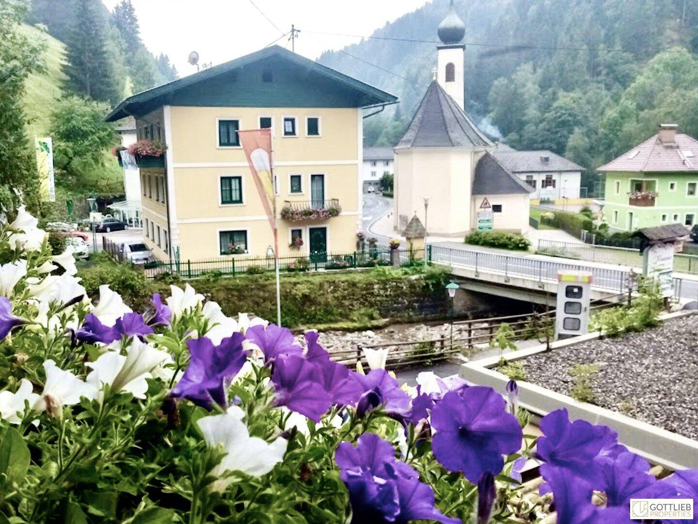 Malerische Ansicht eines Dorfes mit Kirche und Brücke, umgeben von grünen Bergen und Blumen im Vordergrund.