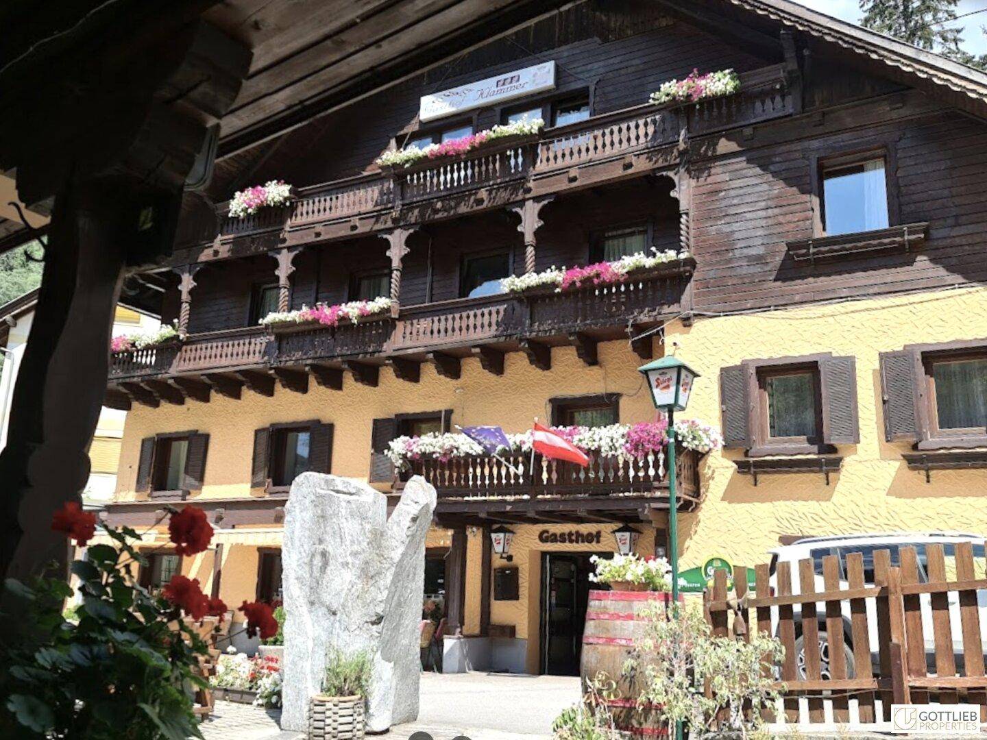 Eingangsbereich eines traditionellen Gasthofs mit Holzbalkonen, Blumen und einem "Gasthof"-Schild.