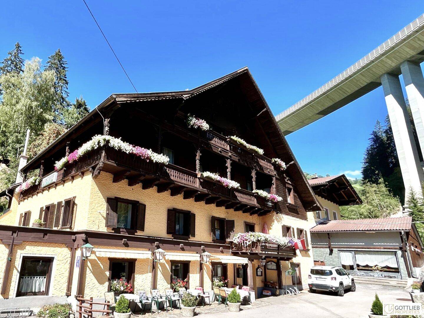Traditionelles Gasthaus mit Holzbalkonen und Blumenkästen unter einer großen Brücke an einem sonnigen Tag.