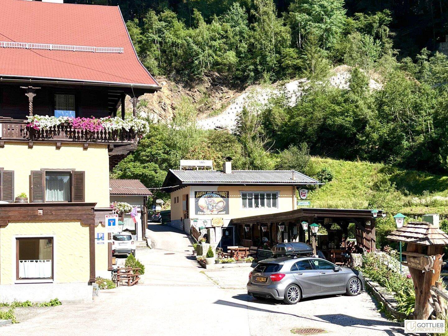 Ansicht einer ländlichen Straße mit traditionellen Gebäuden, geparktem Auto und bewaldeten Hügeln im Hintergrund.