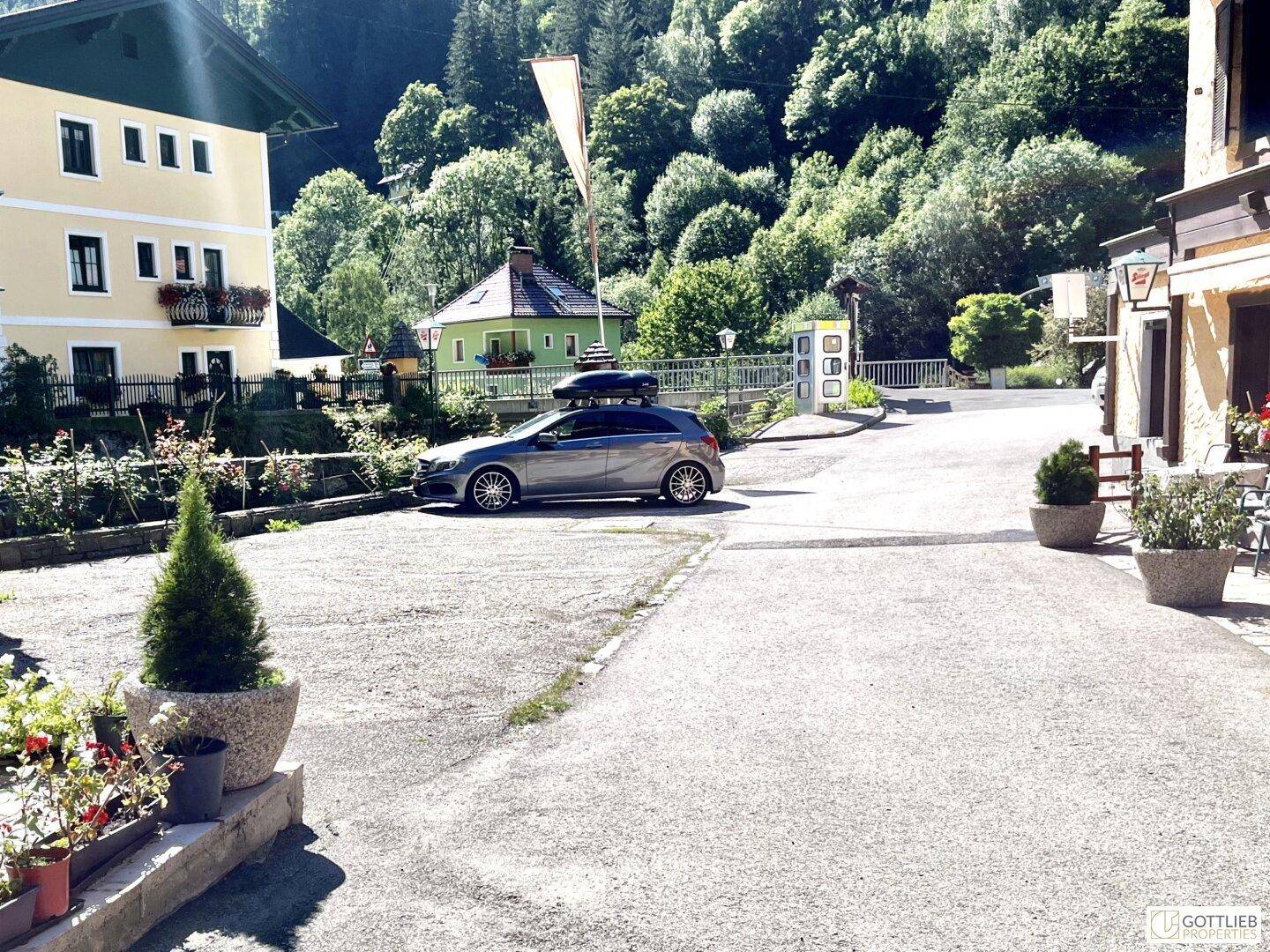 Großer Parkplatz vor traditionellen Gebäuden, umgeben von grünen Bergen unter blauem Himmel.