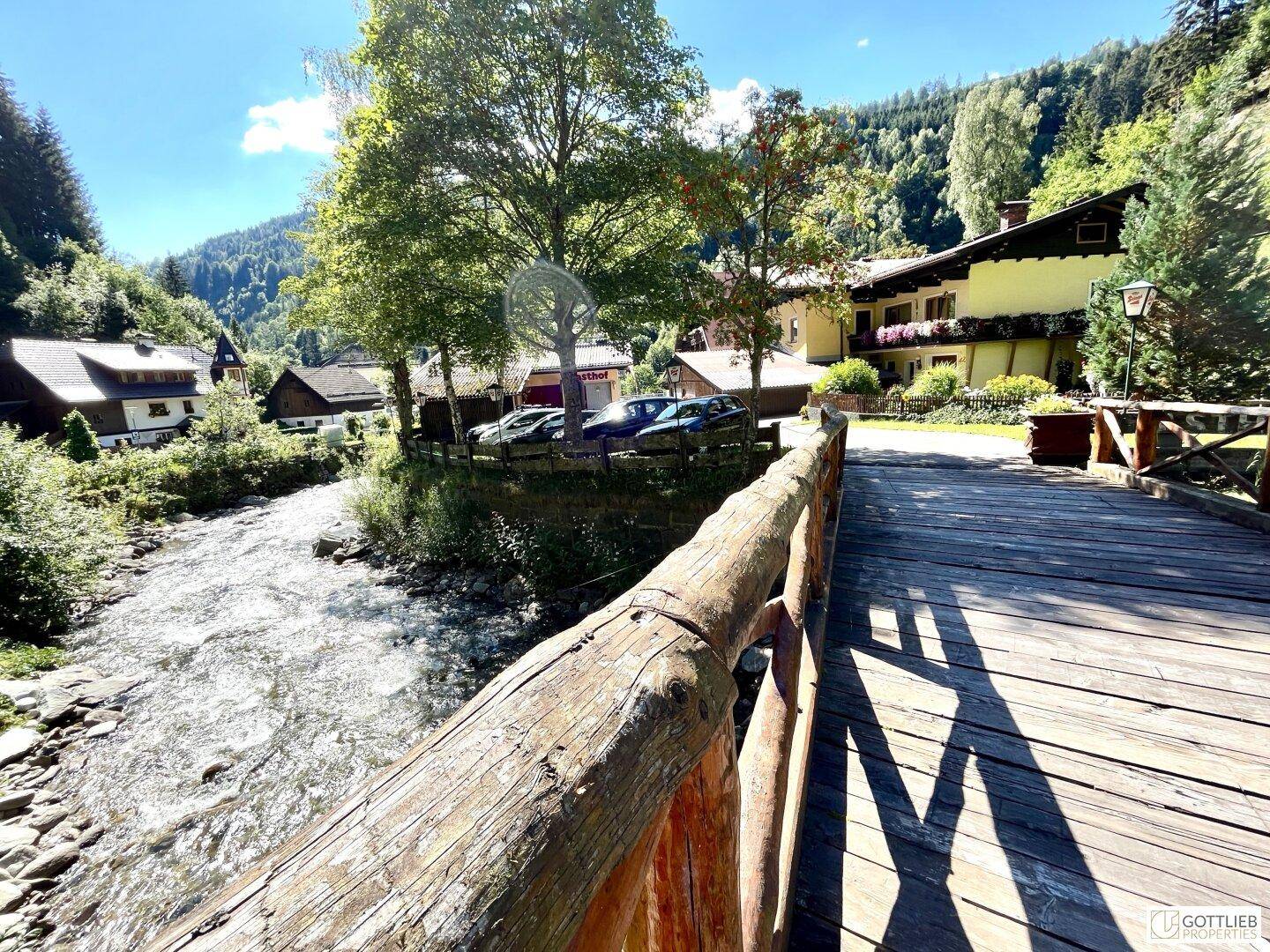 Reißender Fluss mit einer rustikalen Holzbrücke im Vordergrund und traditionellen Gebäuden in der Ferne.