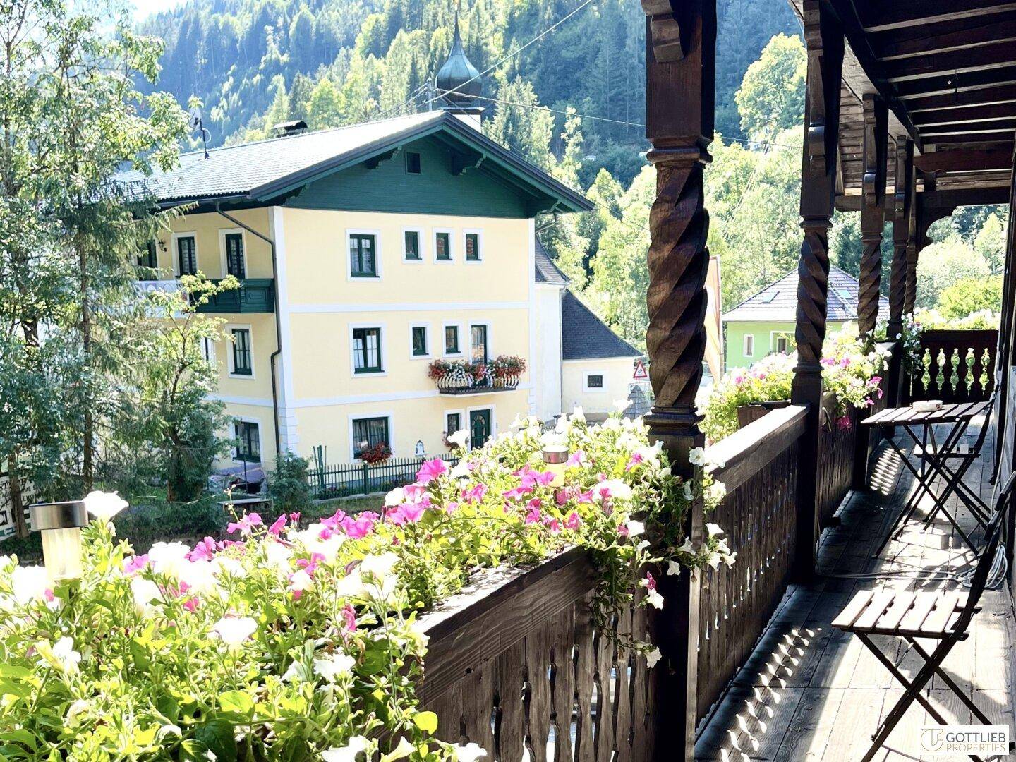 Sonniger Balkon mit bunten Blumenkästen und Blick auf die umliegenden Berge und Gebäude.