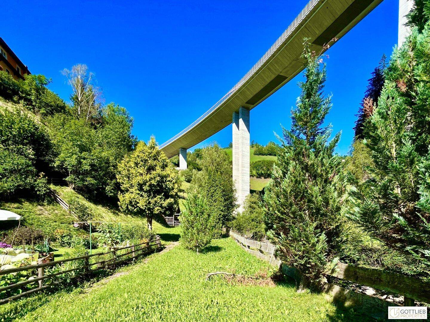 Eine lange, moderne Brücke überspannt ein grünes Tal mit bewaldeten Hängen unter einem strahlend blauen Himmel.