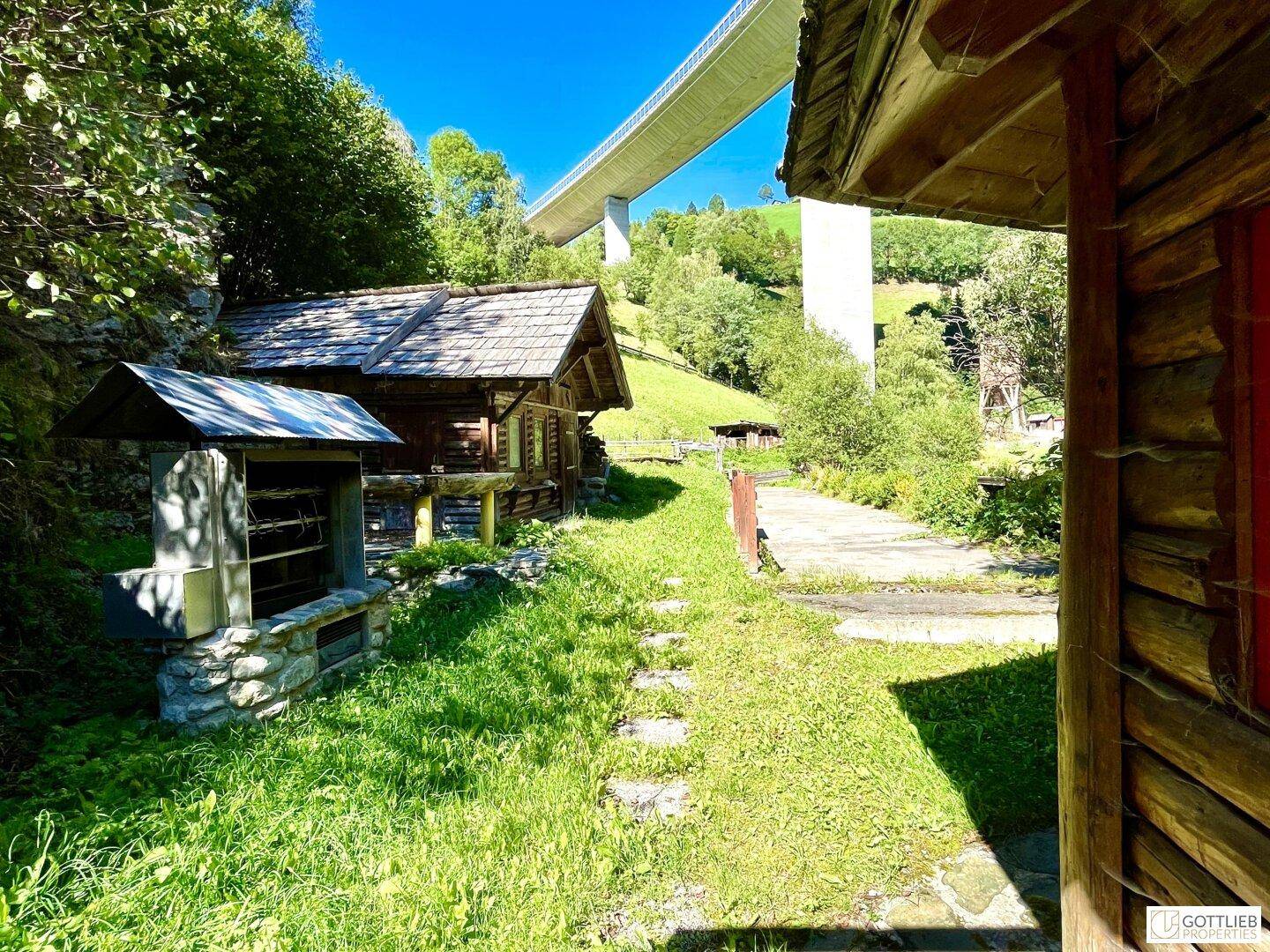 Rustikale Holzhütten und ein Grillplatz im Grünen, mit einer imposanten Brücke im Hintergrund, umgeben von Natur.