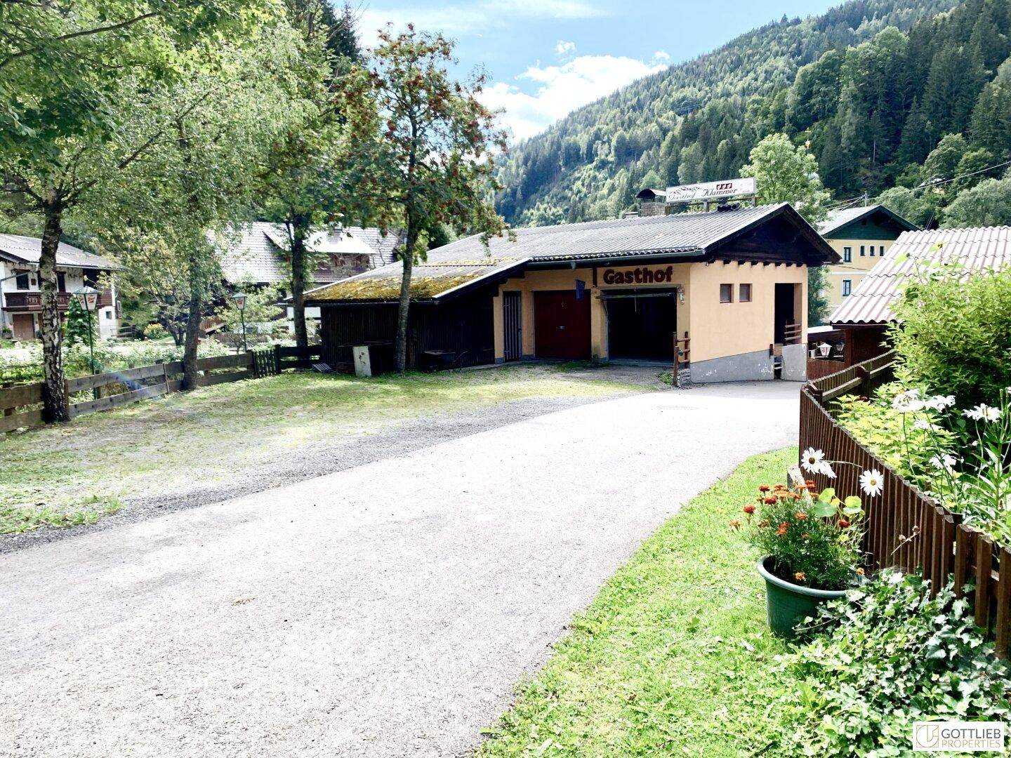 Außenansicht eines Gasthofs mit großem Vorplatz und umliegenden grünen Bergen, eingebettet in die Landschaft.