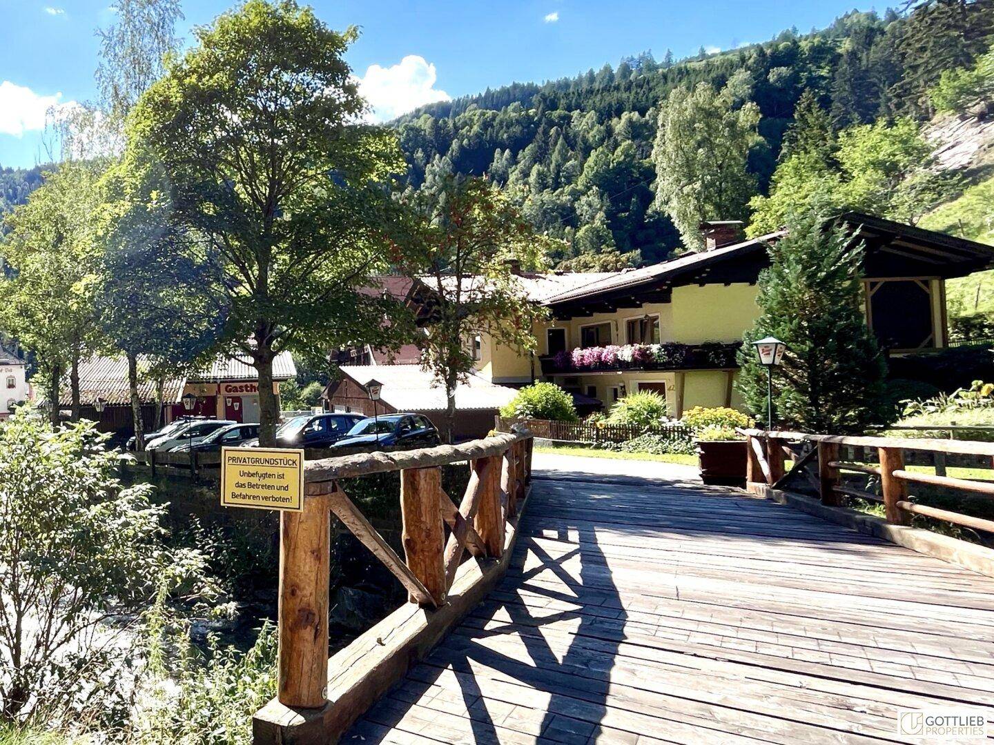 Traditioneller Gasthof mit Holzbalkonen und einer hölzernen Brücke im Vordergrund, umgeben von üppiger Natur.