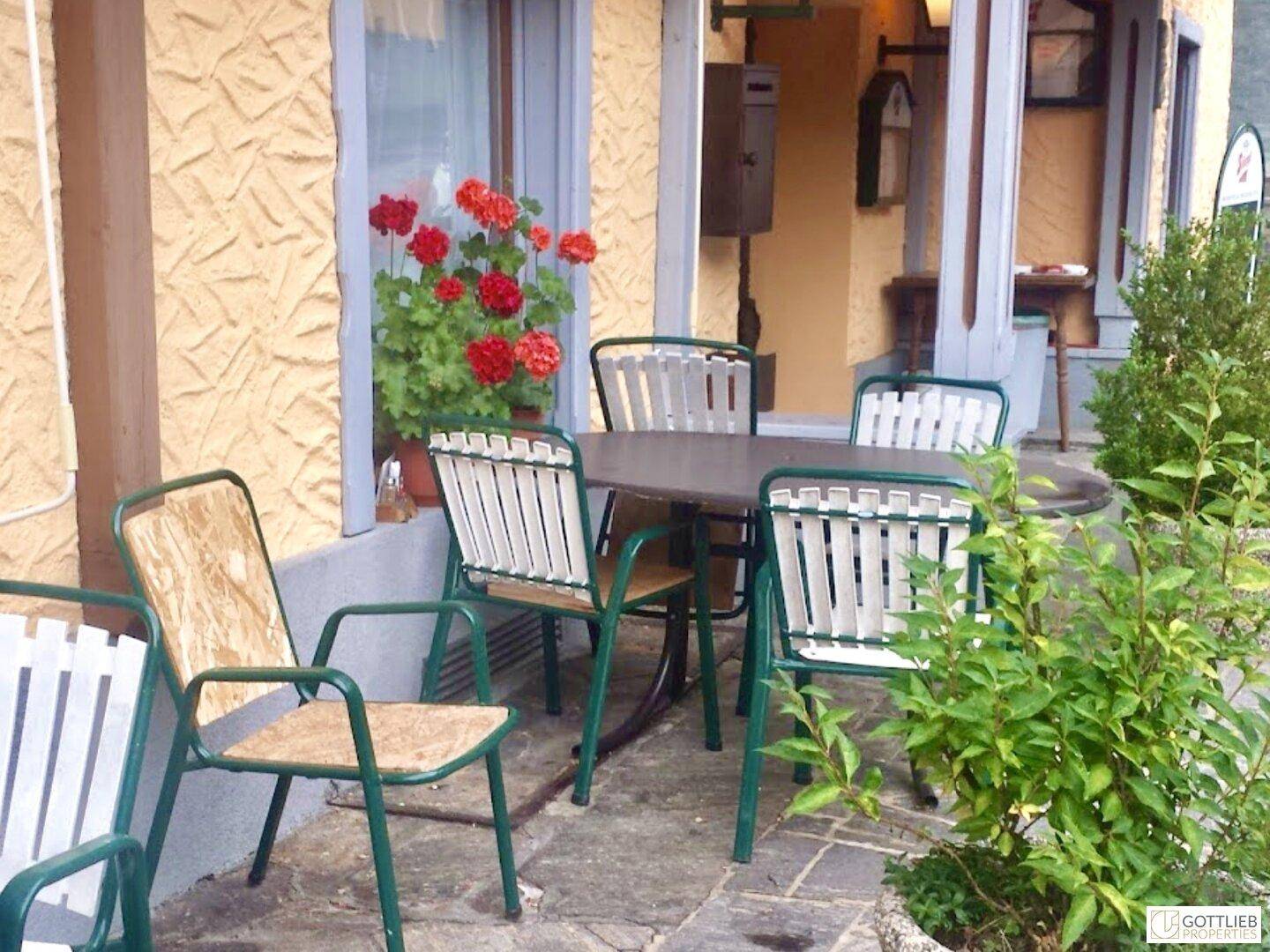 Gemütlicher Außenbereich mit Tischen und Stühlen, dekoriert mit roten Geranien, ideal für entspannte Stunden im Freien.