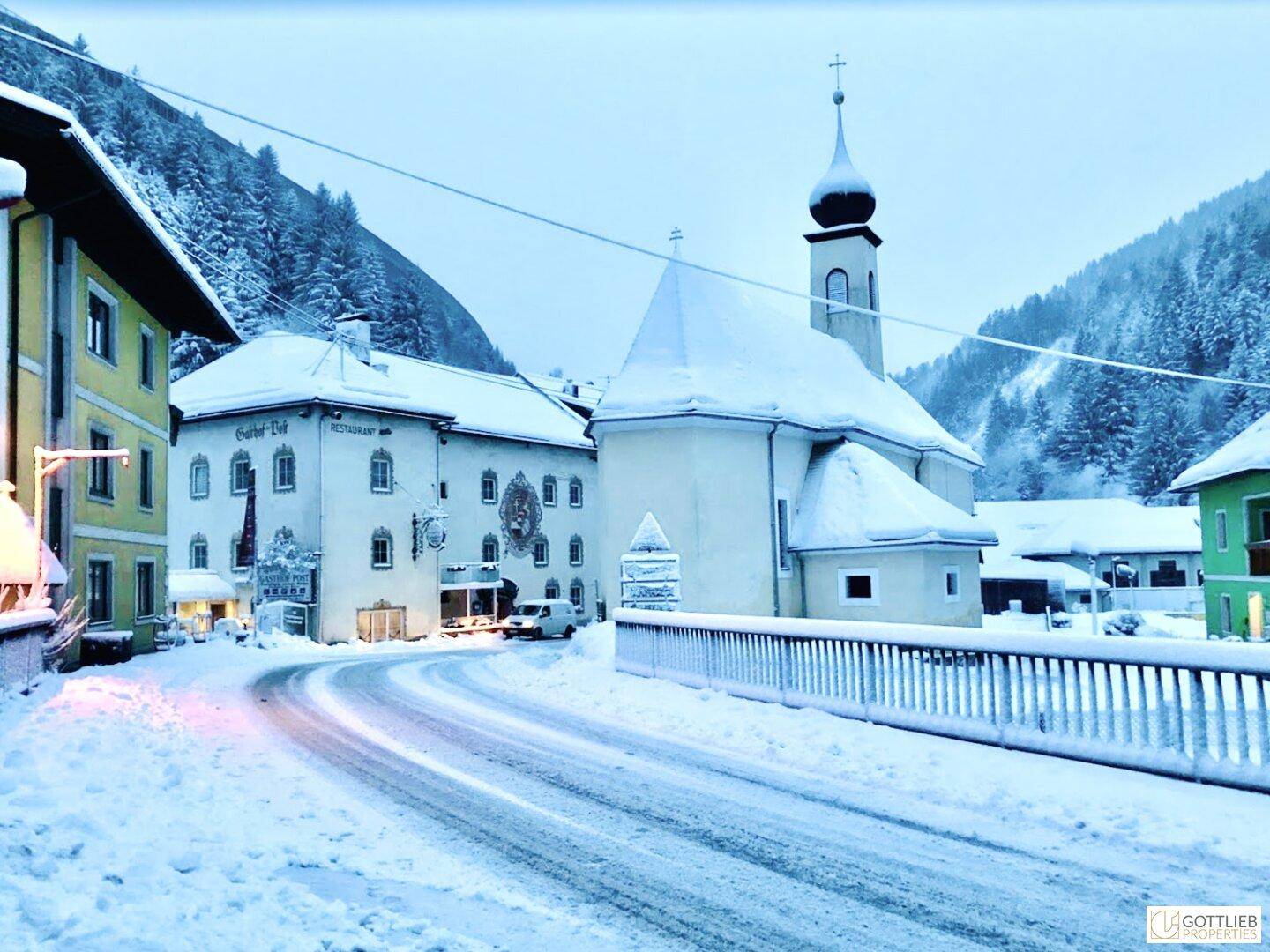 Winterliche Dorfstraße mit verschneiten Häusern und einer Kirche unter einem hellblauen Himmel, eine idyllische Szene.