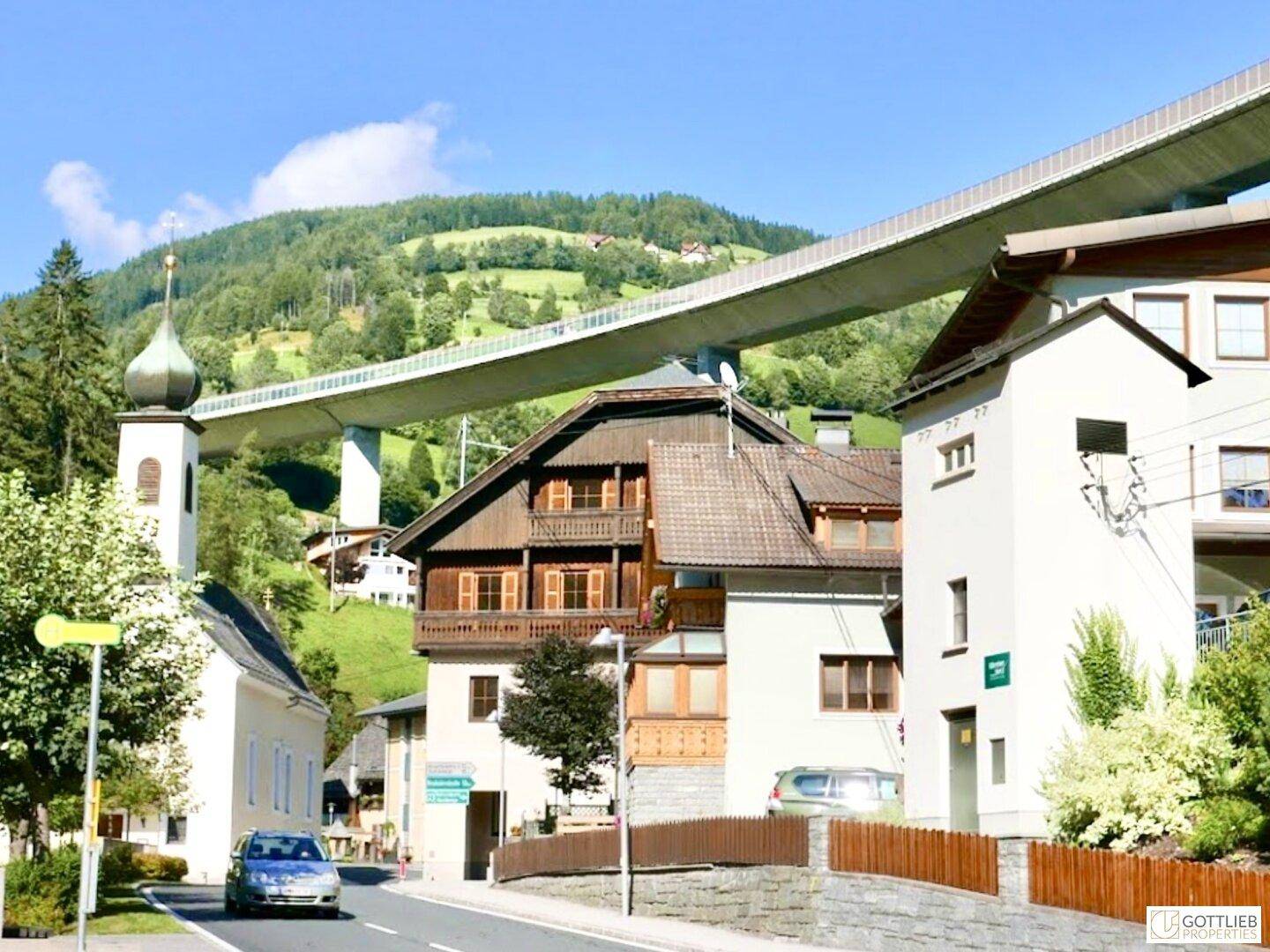 Malerisches Dorf mit einer Kirche und einer großen Brücke im Hintergrund, umgeben von grünen Hügeln und Natur.