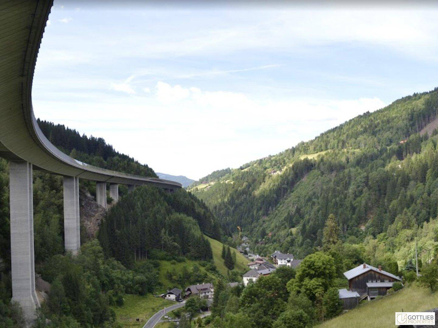 Weitläufiges Tal mit einer imposanten Brücke, umgeben von dichten Wäldern und einem kleinen Dorf, eine beeindruckende Landschaft.