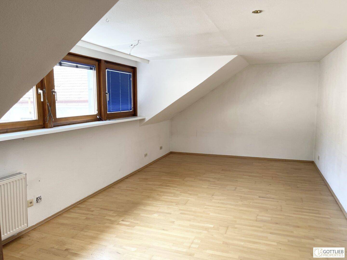Ein heller Raum im Dachgeschoss mit schrägen Wänden, Holzfußboden und einem Heizkörper unter dem Fenster.