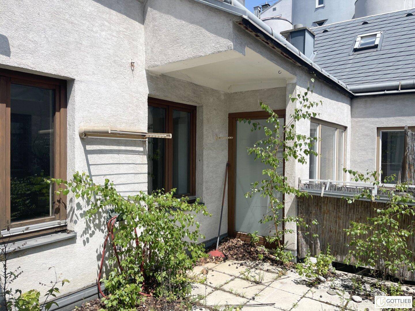 Eingangsbereich eines Gebäudes mit einer kleinen, ungepflegten Terrasse und braunen Fensterrahmen.