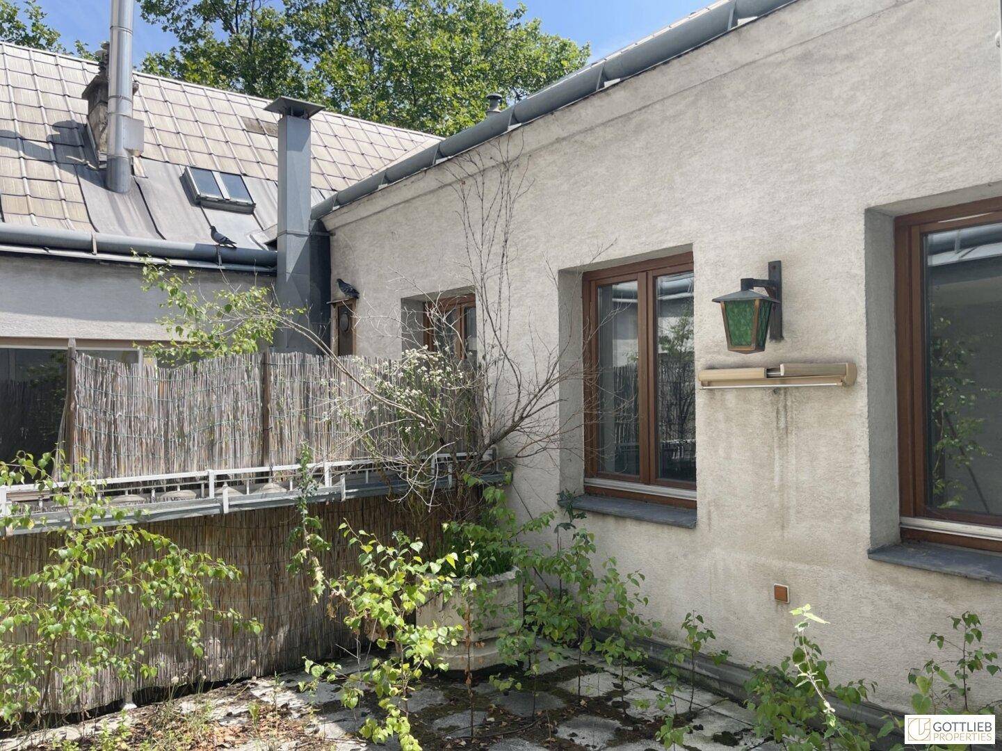 Außenansicht eines Gebäudes mit braunen Fenstern und einer kleinen, bewachsenen Terrasse im Hinterhof.