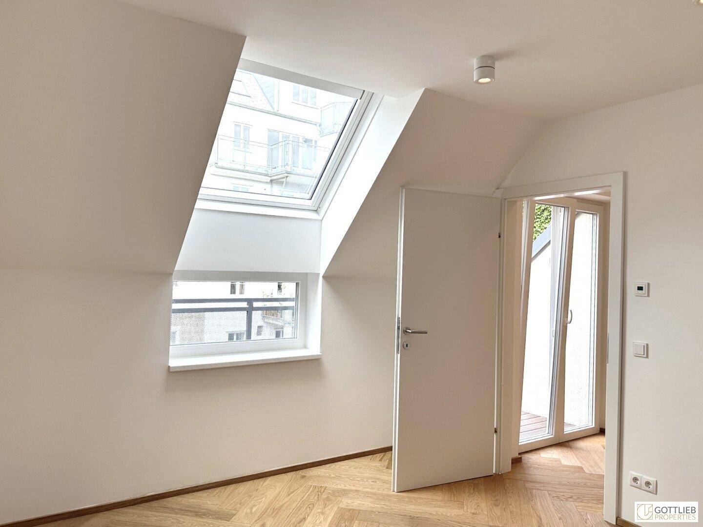 Heller Raum mit Dachschrägen, Fischgrätparkett, Dachfenster und Zugang zu einem Außenbereich.