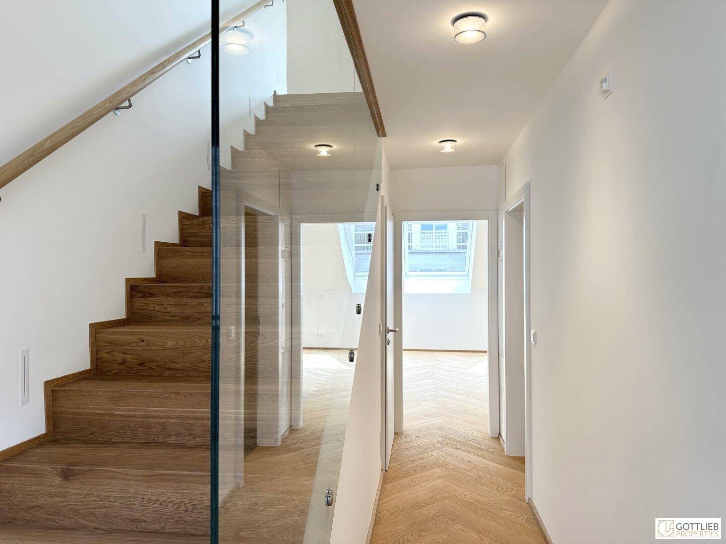 Heller Flur mit Fischgrätparkett und moderner Holztreppe mit Glasgeländer.
