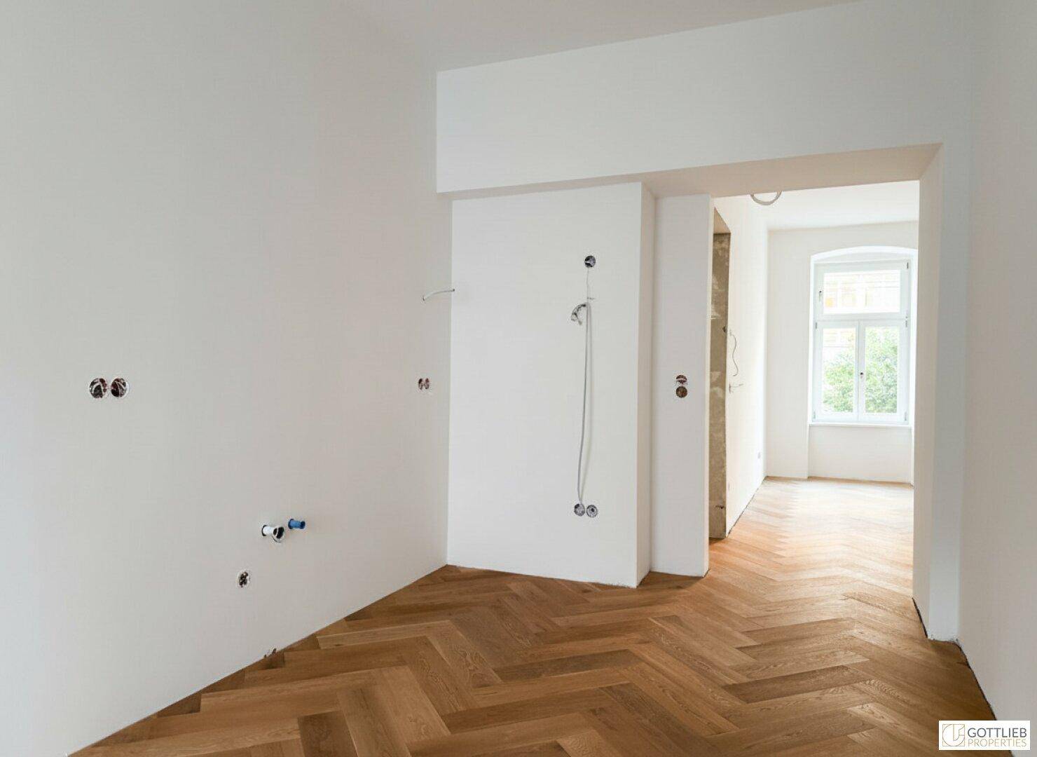 Rohbau einer zukünftigen Küche mit Fischgrätparkett und vorbereiteten Anschlüssen.