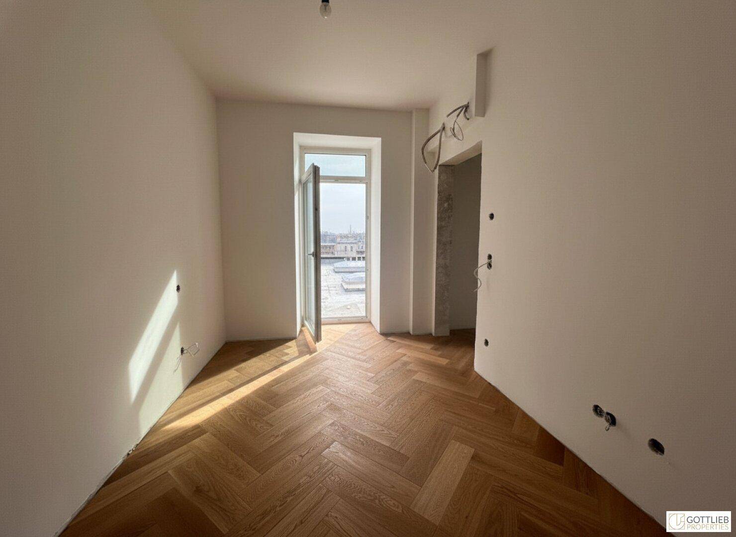 Geräumiges Zimmer mit Fischgrätparkett und offener Balkontür, die den Raum belichtet.