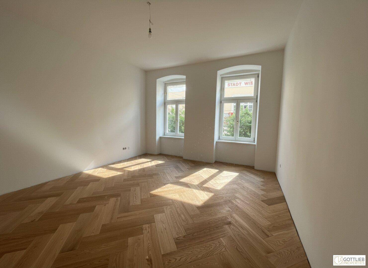 Heller Raum mit Fischgrätparkett und zwei Fenstern, die den Raum mit natürlichem Licht füllen.