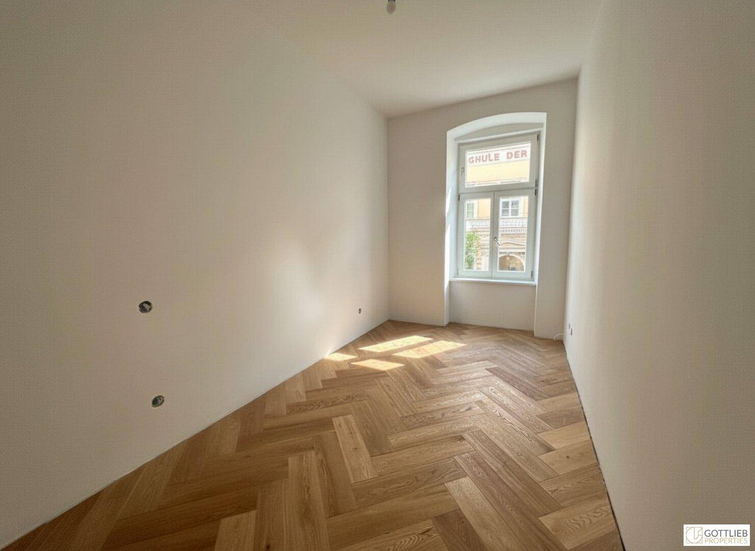 Helles Schlafzimmer mit Fischgrätparkett und einem Fenster, das viel Sonnenlicht hereinlässt.
