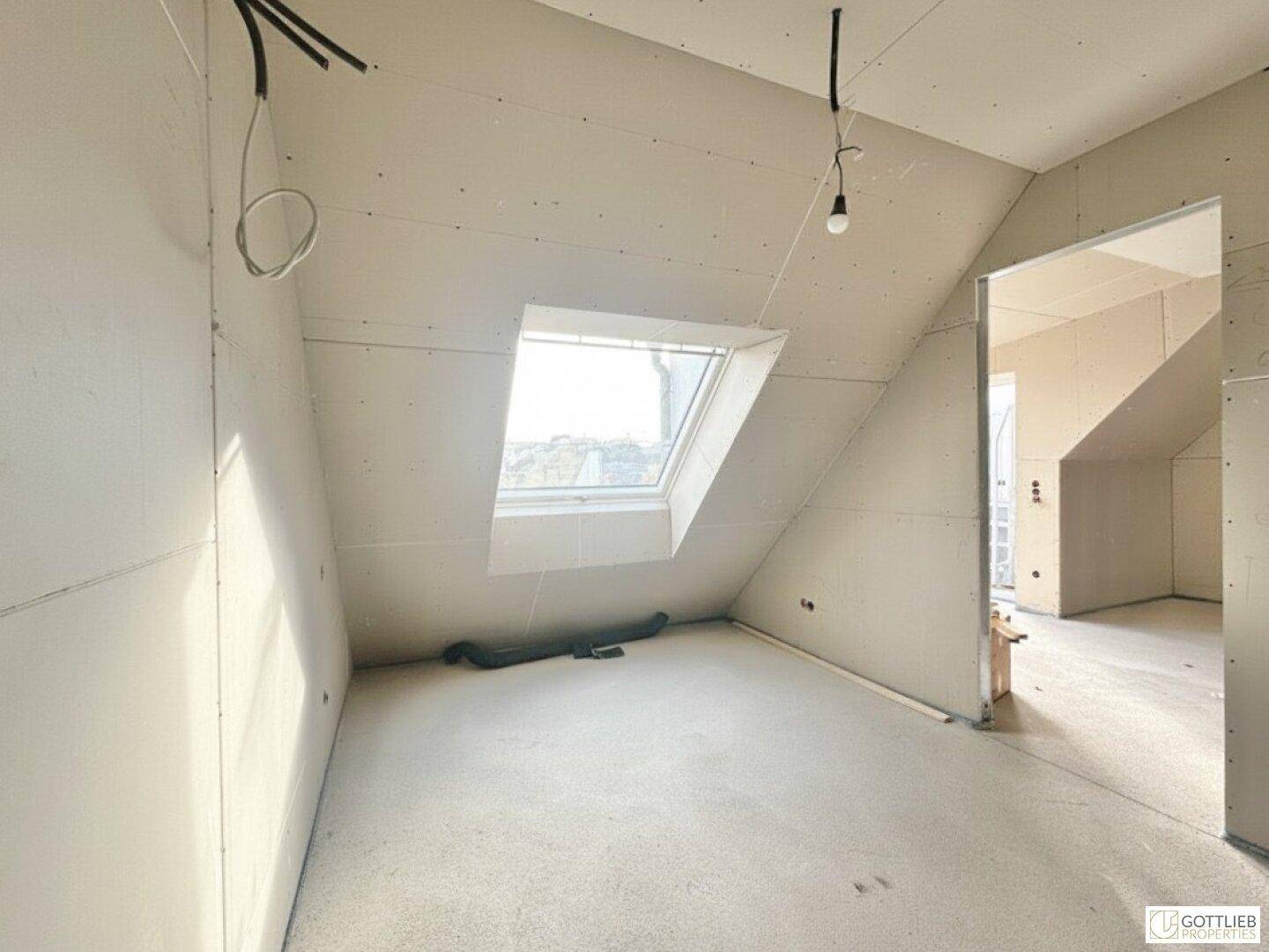 Unfertiger Raum mit schräger Decke und einem einzelnen Dachfenster, das Licht hereinlässt.