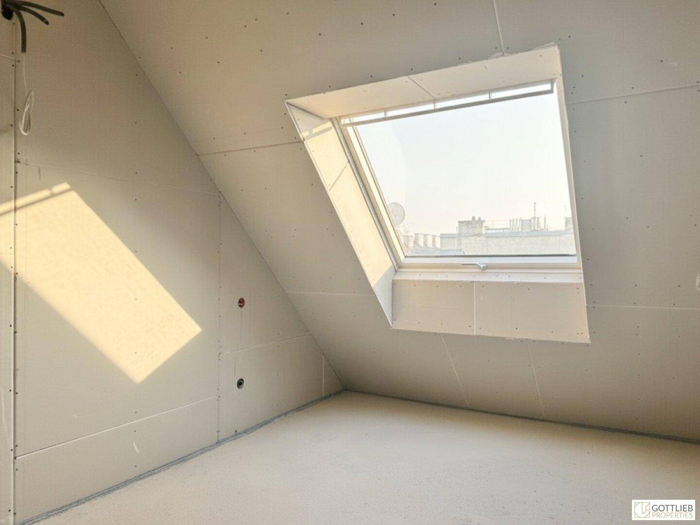 Unfertiger Raum mit schräger Decke und einem großen Dachfenster, das viel Licht spendet.