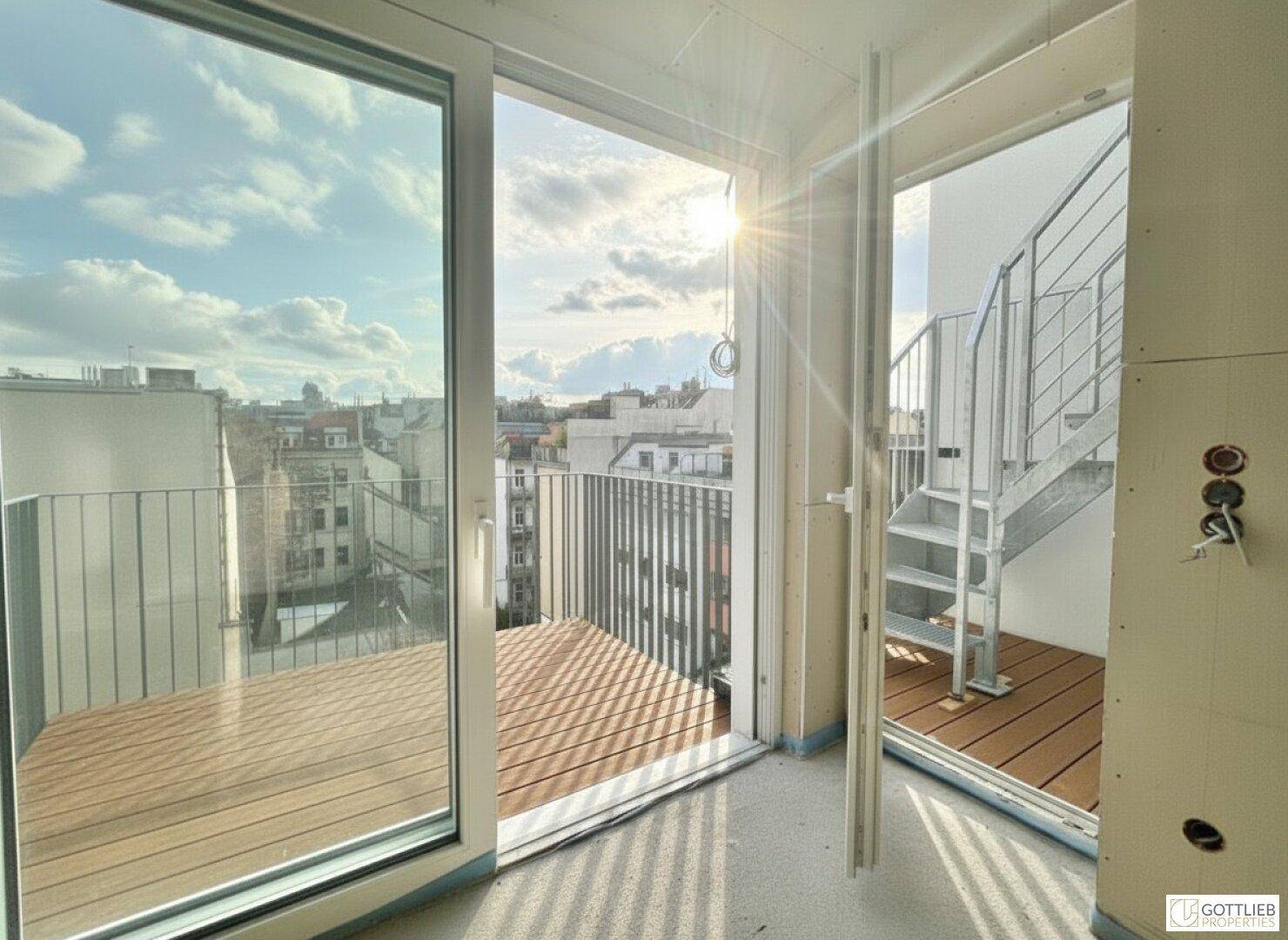 Innenraum mit Zugang zu einem Balkon mit Holzbelag und Außentreppe unter strahlendem Himmel.