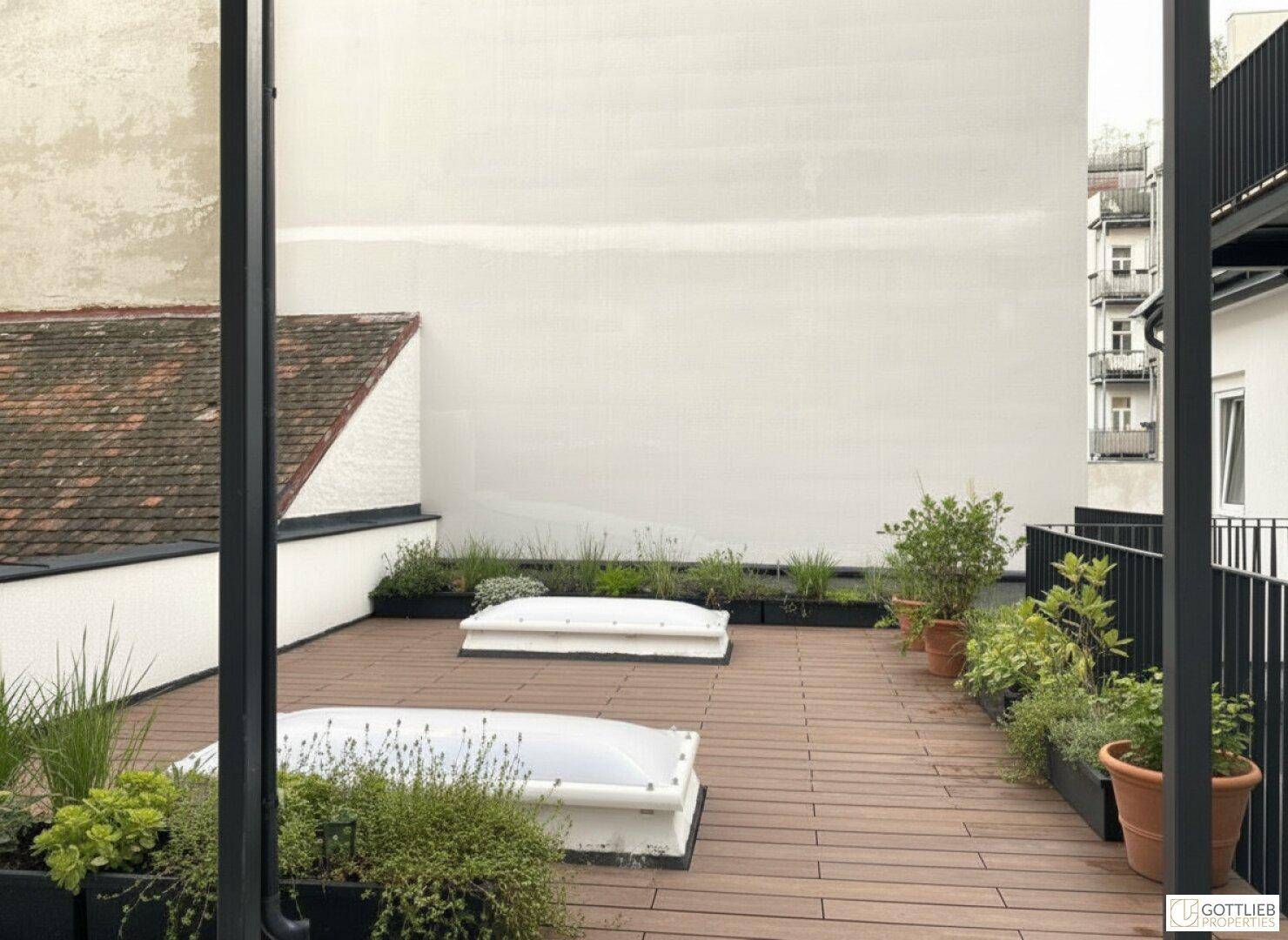Geräumige Dachterrasse mit Holzbelag und grünen Pflanzen in Kübeln, umgeben von Gebäuden.