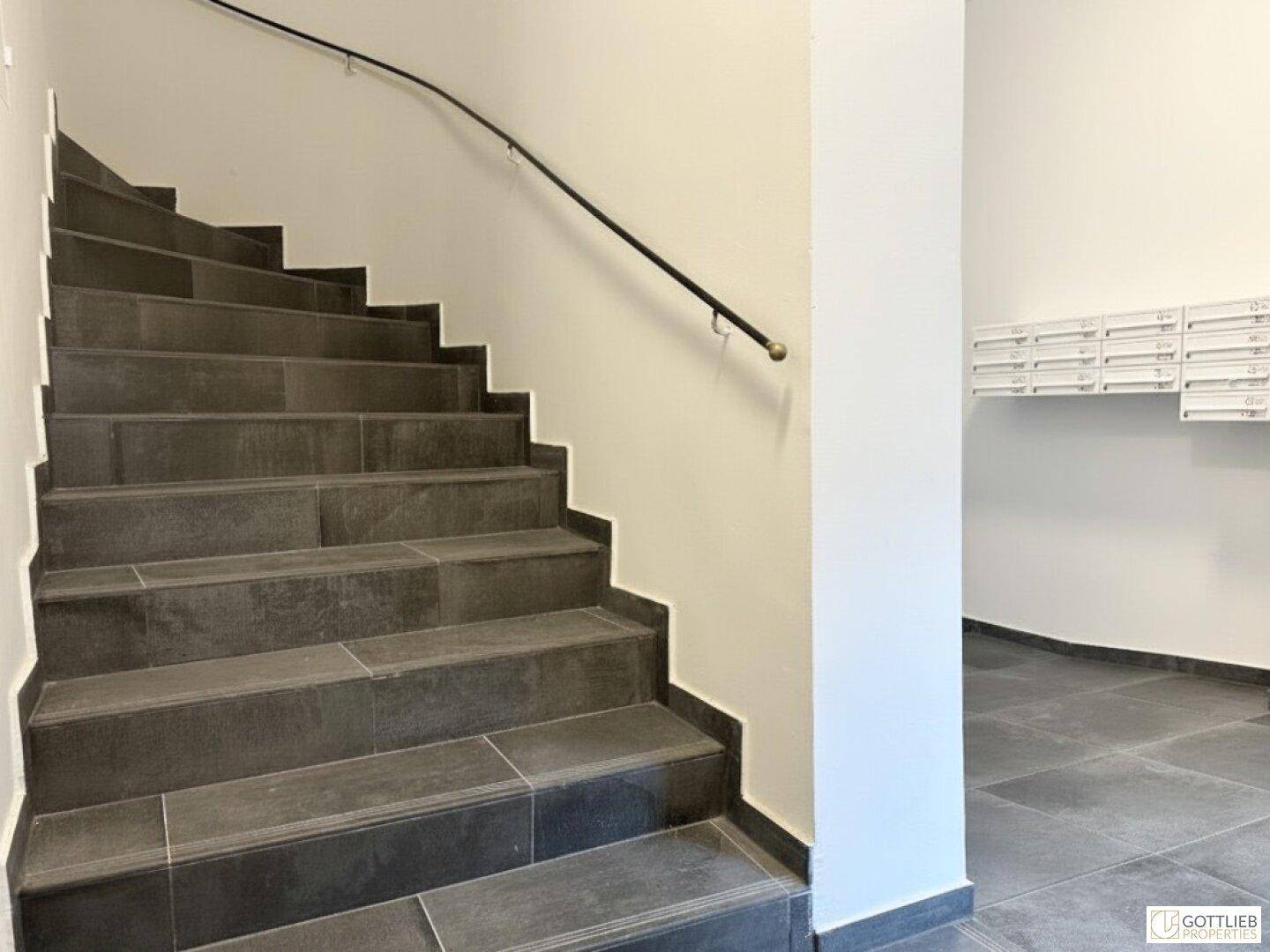 Treppenhaus mit dunklen Fliesenstufen und schlichtem Geländer, führt zu den oberen Etagen.