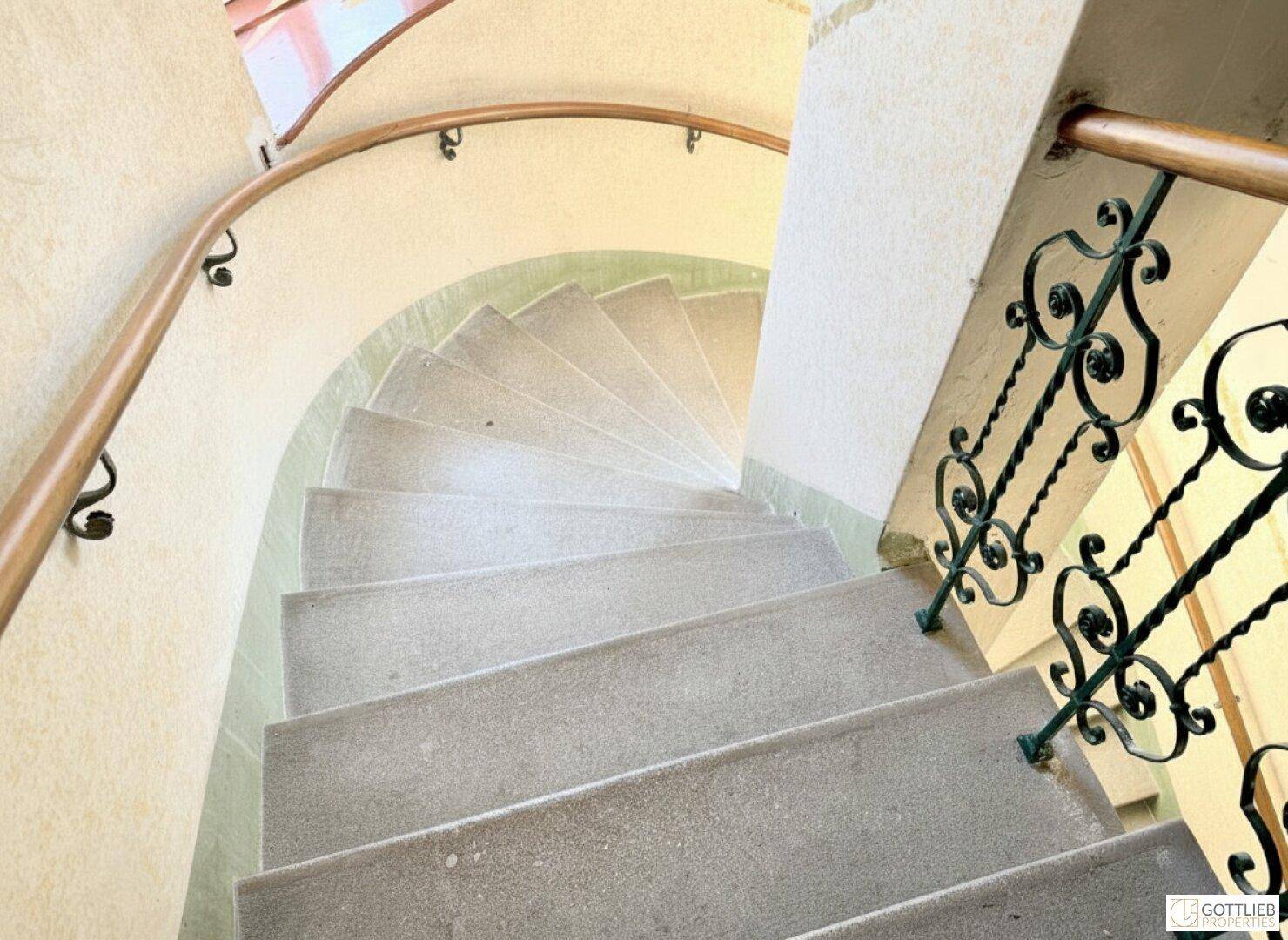 Eine geschwungene Steintreppe mit schmiedeeisernem Geländer und Holzhandlauf in einem Altbau.