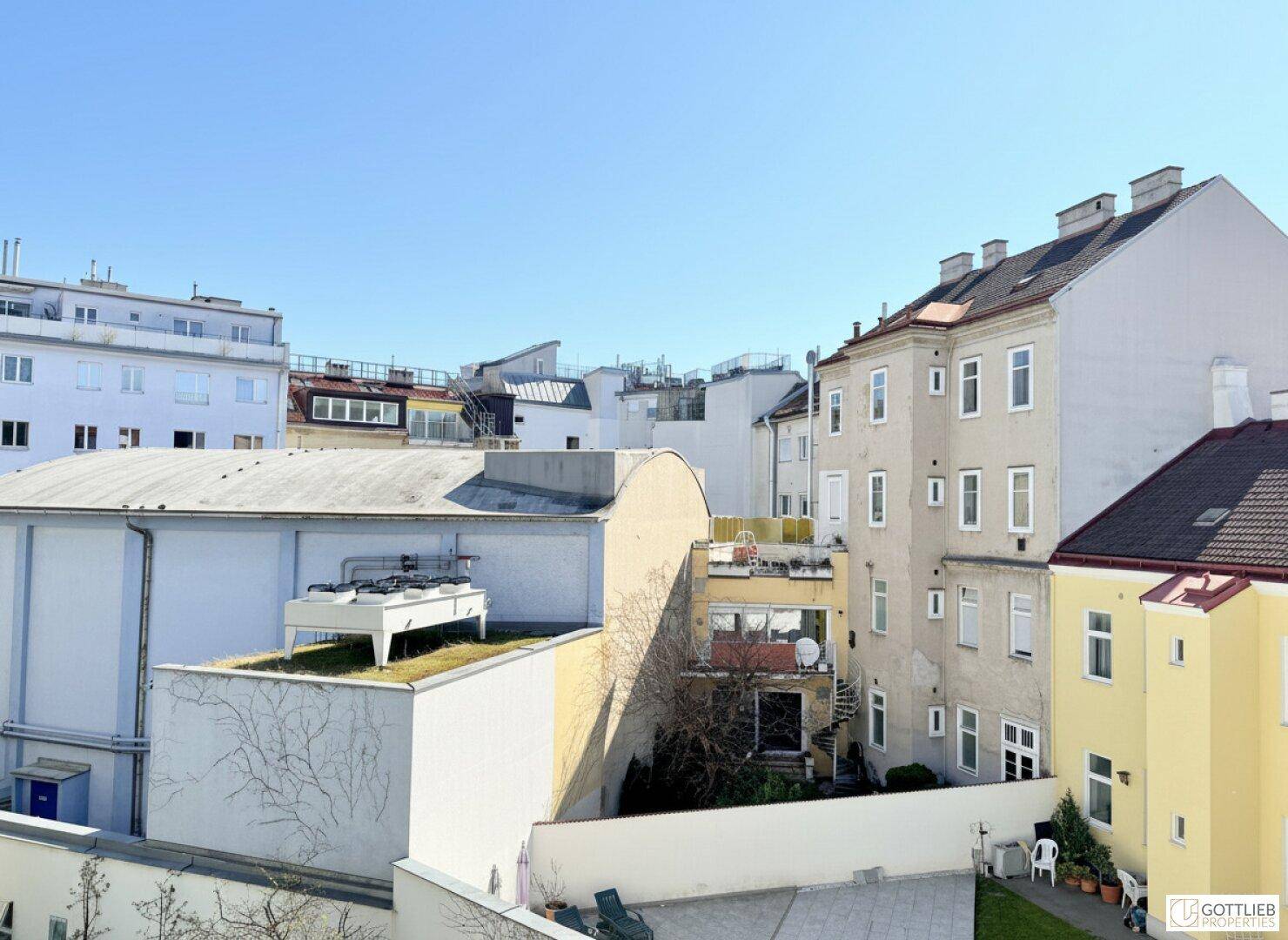 Blick über einen Innenhof mit verschiedenen Gebäuden und Dächern unter klarem, blauem Himmel.