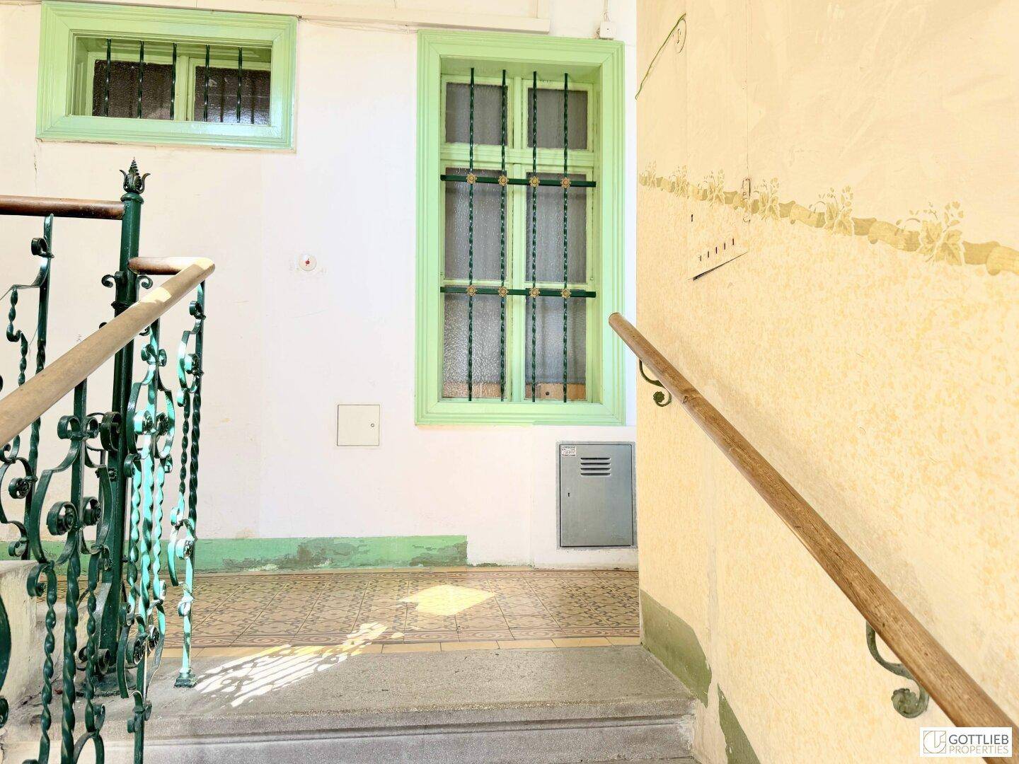 Treppenabsatz mit gemusterten Fliesen, hellgrünen Fensterrahmen und einem schmiedeeisernen Geländer.