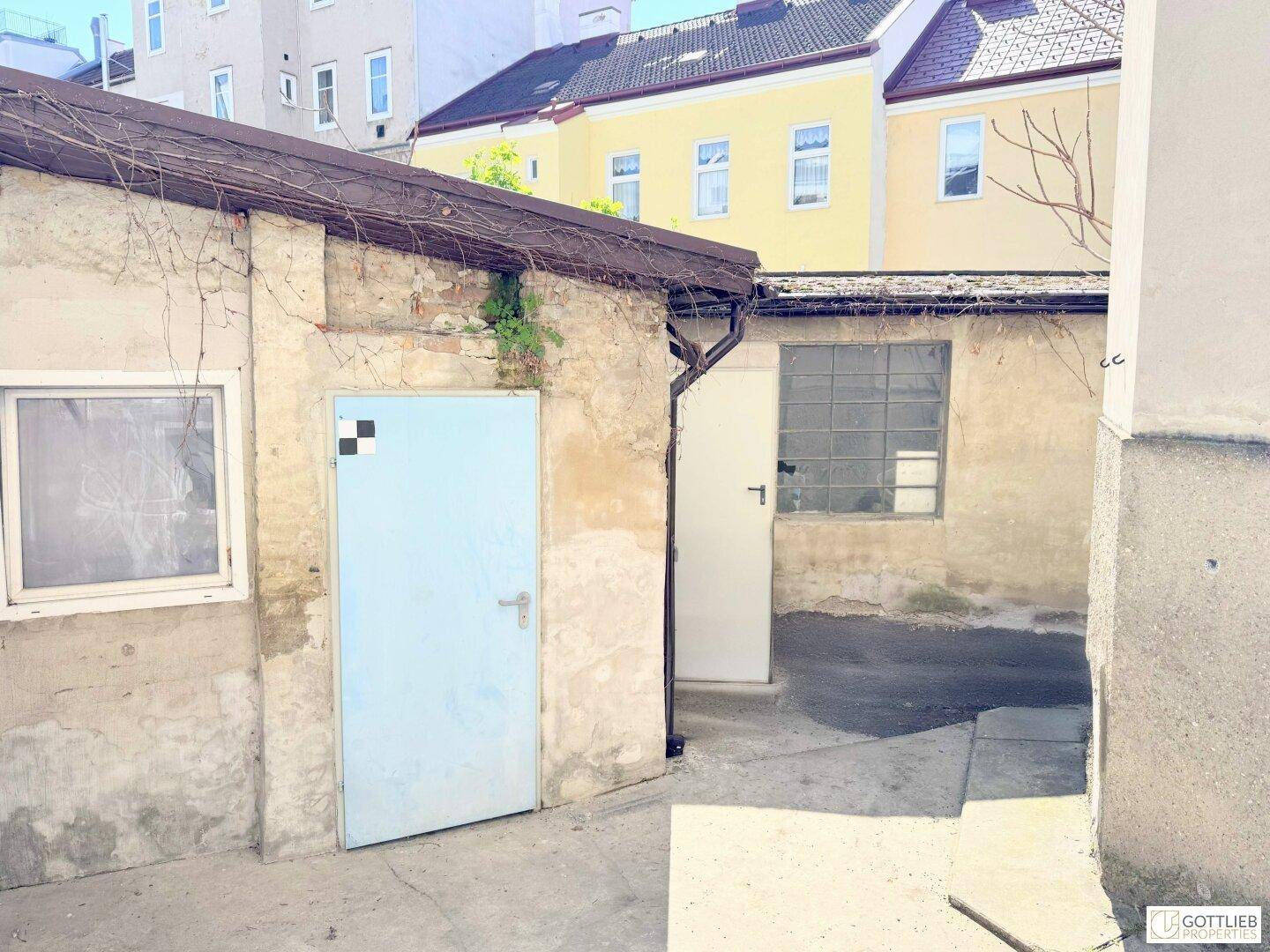 Hinterhofbereich mit zwei kleinen Nebengebäuden, einer blauen Tür und einem Fenster mit Glasbausteinen.
