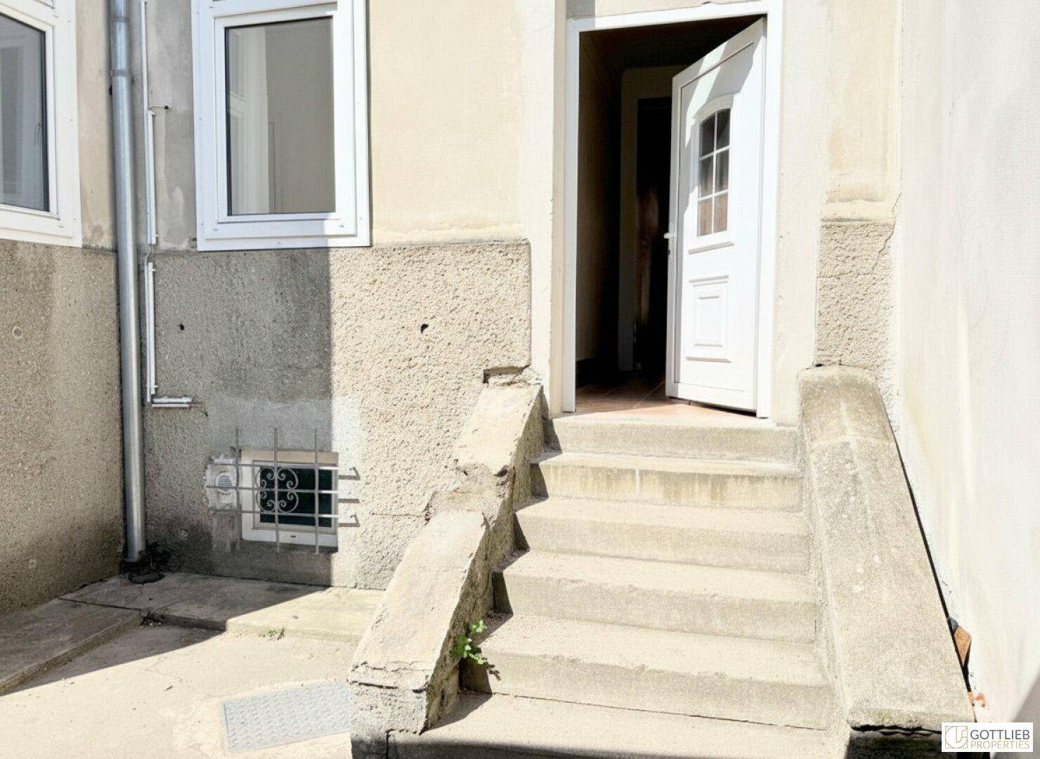 Eingangsbereich mit Steinstufen, einer offenen weißen Tür und einem Fenster an der Hauswand.