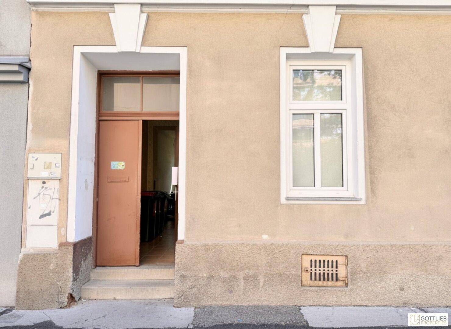 Eingangsbereich eines Gebäudes mit einer geöffneten braunen Tür und einem Fenster daneben.