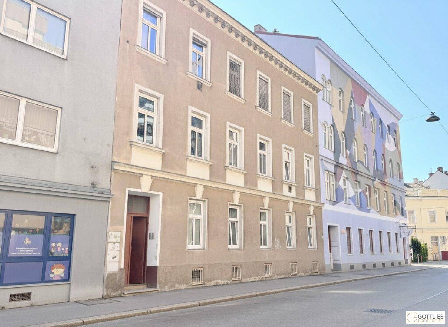 Ansicht eines Wohngebäudes an einer Straße mit weiteren Häusern und blauem Himmel im Hintergrund.