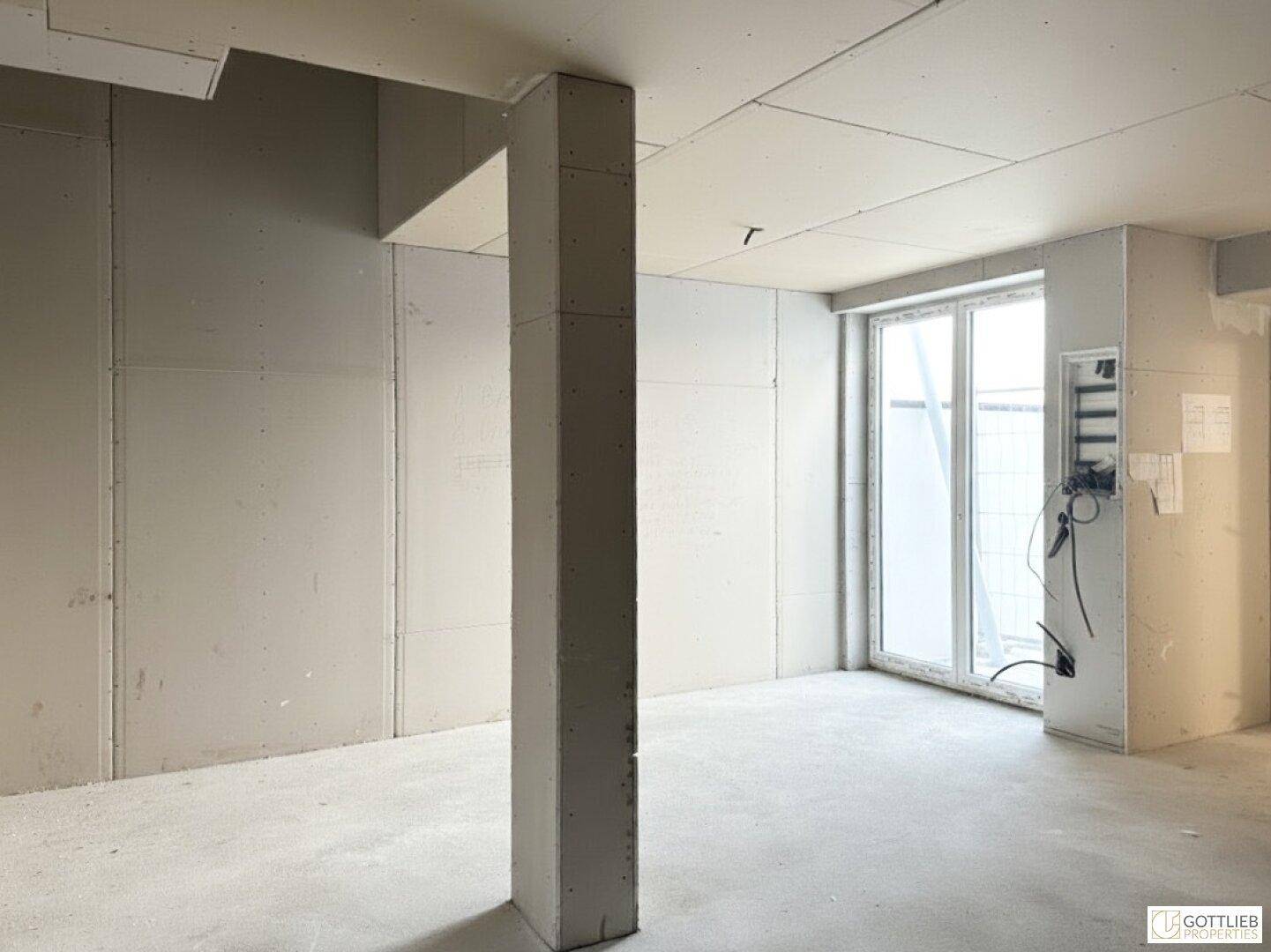Unfertiger Raum mit Trockenbauwänden, Betonboden und großer Fensterfront, noch im Rohbau.