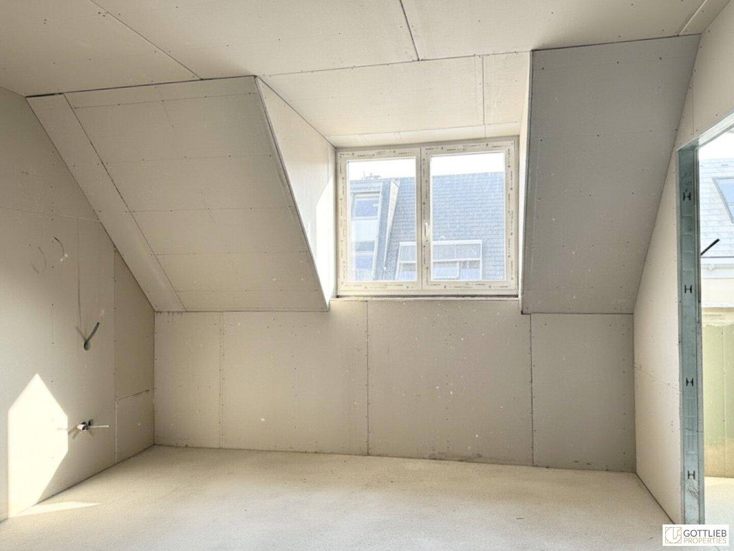 Unfertiger Dachraum mit Trockenbauwänden, Betonboden und Fenster, bereit für den Ausbau.