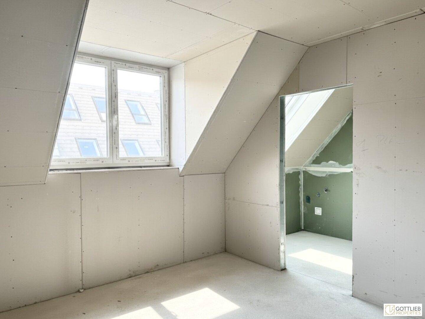Unfertiger Dachraum mit Trockenbauwänden, Betonboden und Fenster, noch im Rohbauzustand.