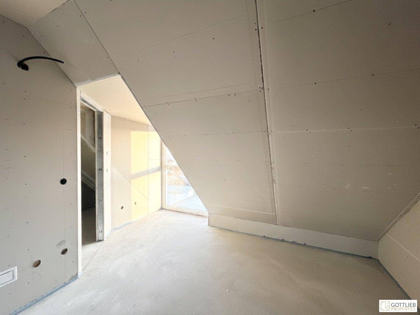 Unfertiger Dachraum mit Trockenbauwänden, Betonboden und schräger Decke, noch im Rohbau.