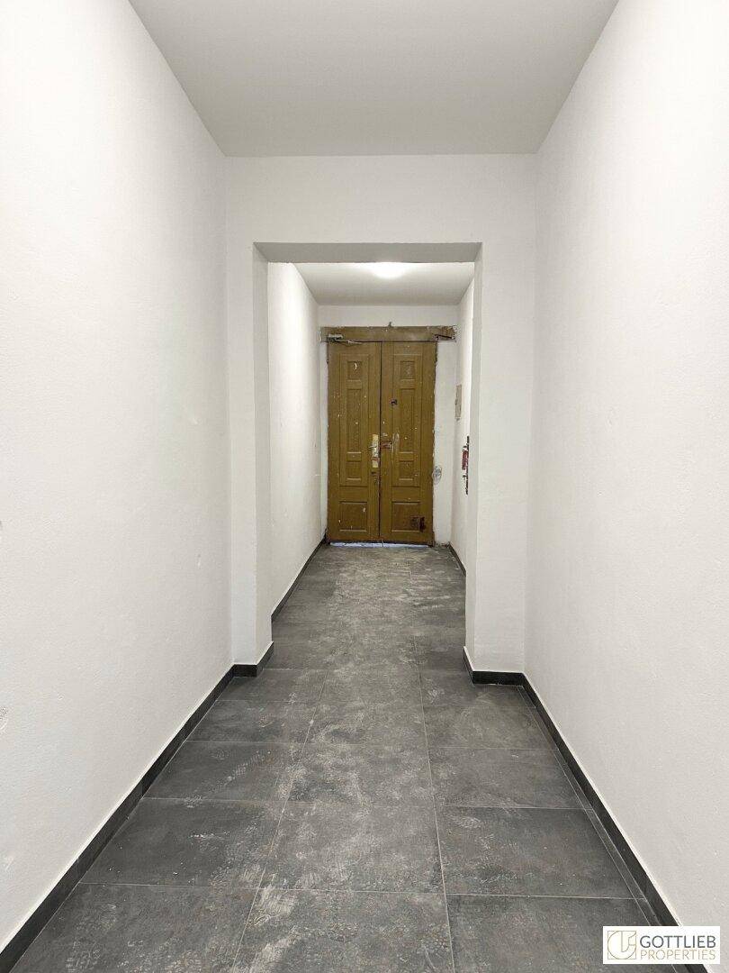 Heller Flur mit dunklem Fliesenboden und weiß gestrichenen Wänden, führt zu einer Holztür.