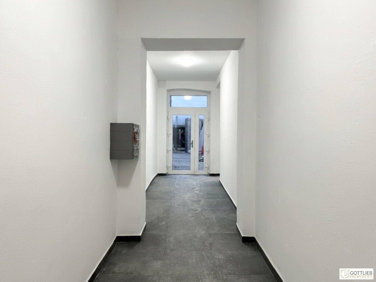 Heller Flur mit dunklem Fliesenboden und weiß gestrichenen Wänden, führt zu einer Glastür.