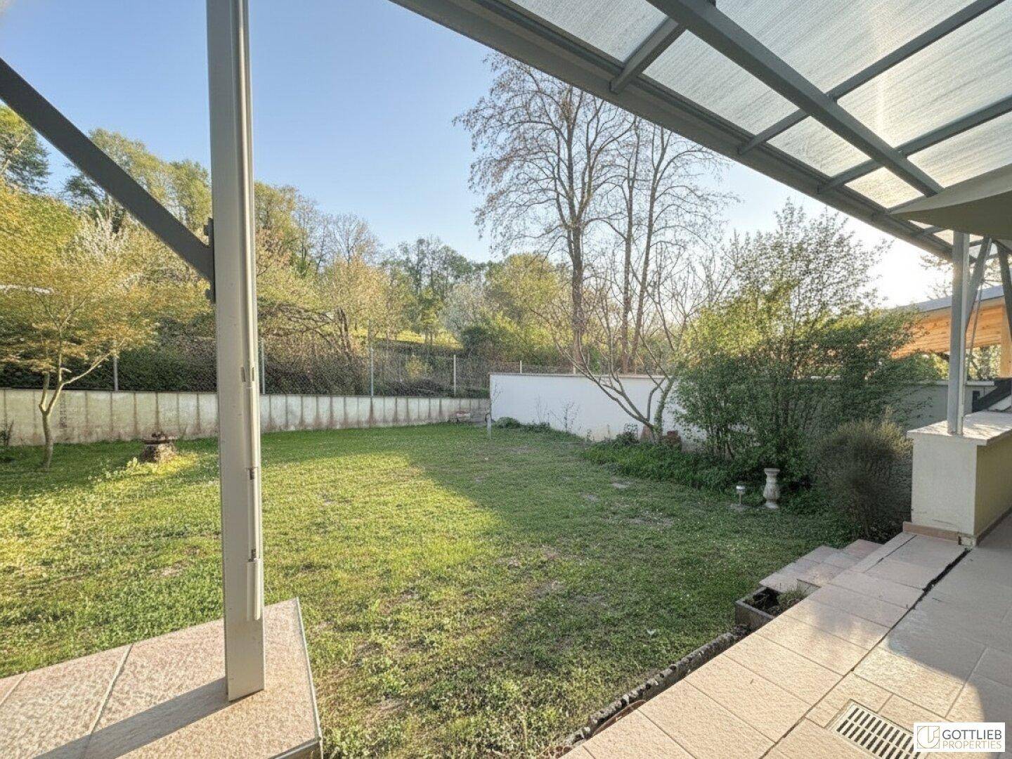 Sonniger Gartenbereich mit Rasenfläche und Bäumen, gesehen von der überdachten Terrasse.