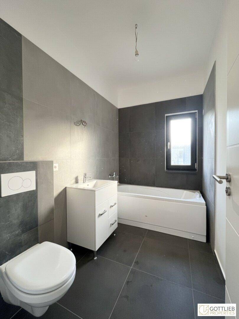Modernes Badezimmer mit grauen Fliesen, Badewanne, WC und einem Fenster für Tageslicht.