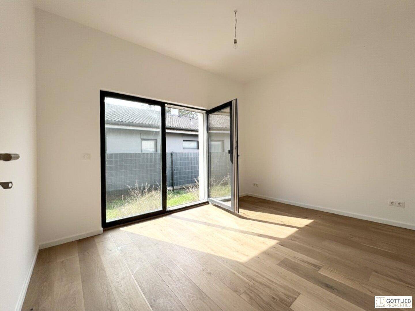 Lichtdurchflutetes Zimmer mit Holzboden und großen Fenstern, die den Zugang zum Außenbereich ermöglichen.