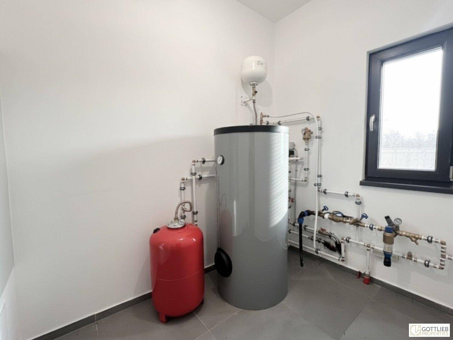 Technikraum mit Warmwasserspeicher, Rohrleitungen und einem Fenster für Belüftung.