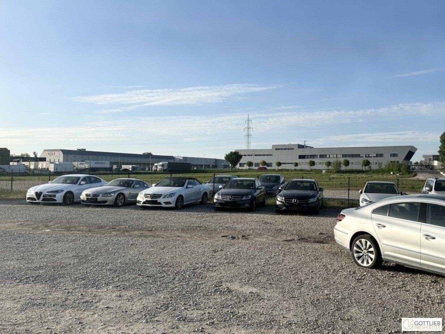 Großer Schotterparkplatz mit geparkten Autos und modernen Industriegebäuden im Hintergrund unter blauem Himmel.